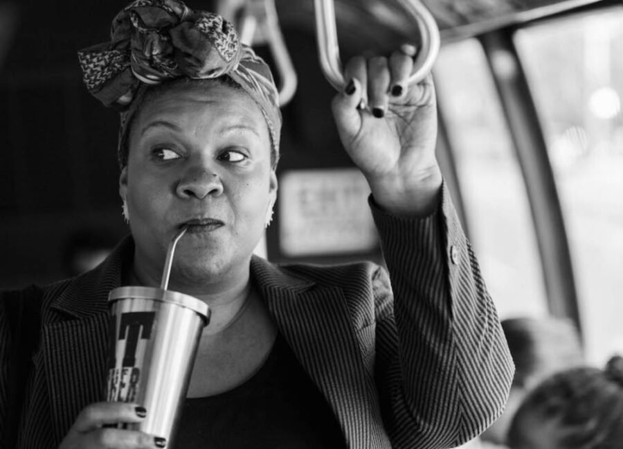 Mulher negra gorda tomando em um copo com canudo, usando um lenço na cabeça e segurando em um ônibus. Está de frente para a câmera e tem os olhos no canto, à direita