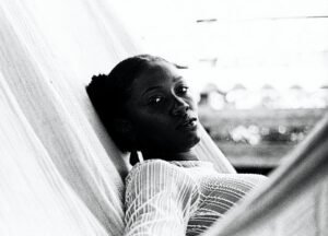 Foto em preto e branco de mulher negra deitada em uma rede olhando para a foto, com rosto sério, porém tranquilo. Usa uma blusa branca de renda de manga comprida.