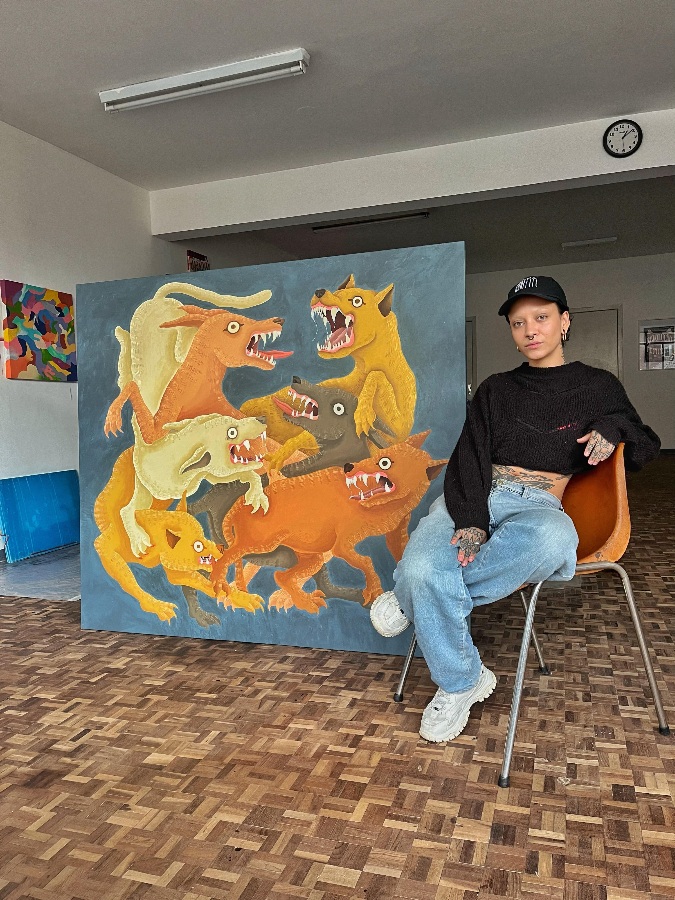 Bruna Pereira posando ao lado de sua obra denominada como Rinha, a técnica utilizada foi: acrílica sobre tela, e se enquadra na arte naif. 